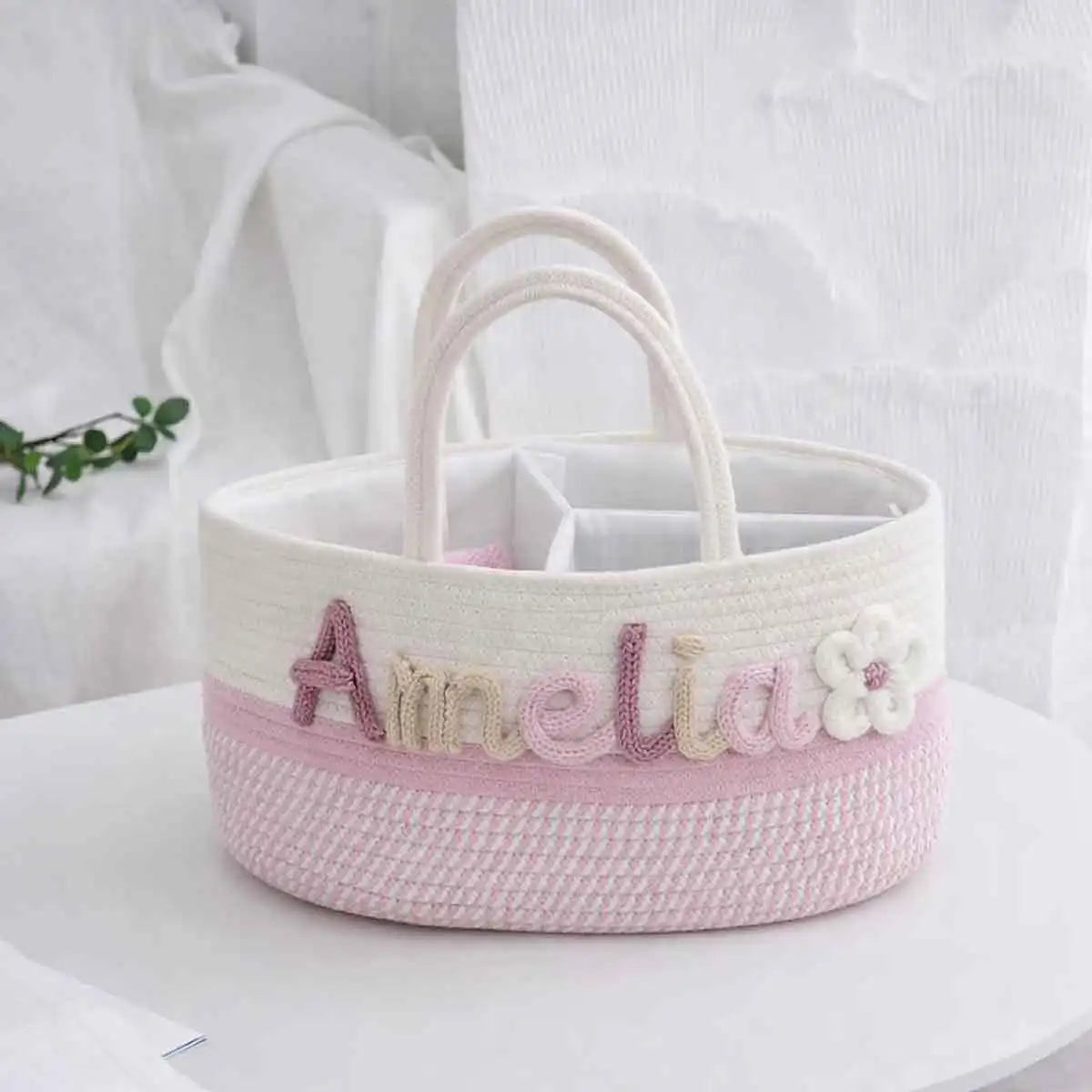 A round woven storage basket with cream handles, a cream top band, and a pink lower band. The front features the name 'Amelia' in embroidered pink and cream letters alongside a white flower applique.