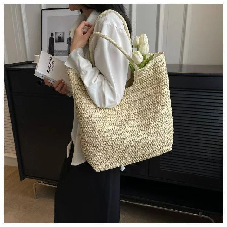 A woman wearing a white satin shirt and black skirt carries a large woven straw tote bag with white tulips tucked inside, standing in front of a black cabinet.