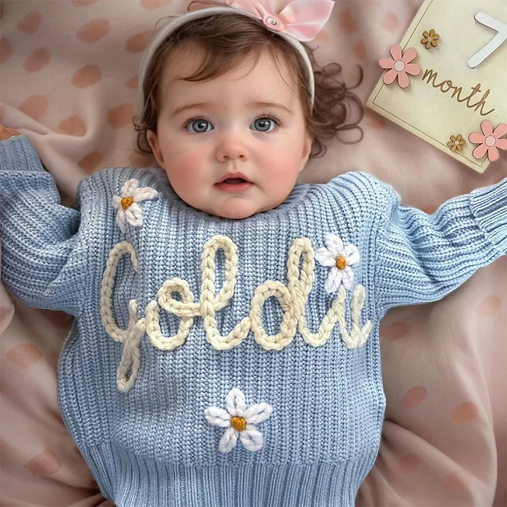 Baby wearing a blue sweater with 'Goldie' embroidered on it, lying on a pink polka dot blanket.