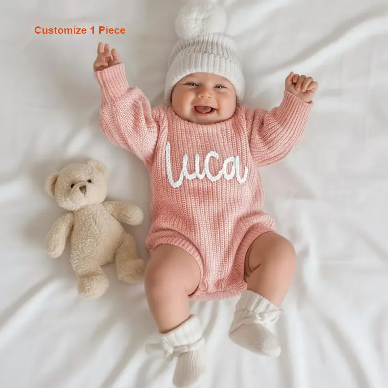 A baby lying on white bedding wearing a pink ribbed knit one-piece with the name 'Luca' in white script, a white knit pom-pom hat, and white booties. A beige teddy bear sits beside the baby, and the text 'Customize 1 Piece' appears in the top left.