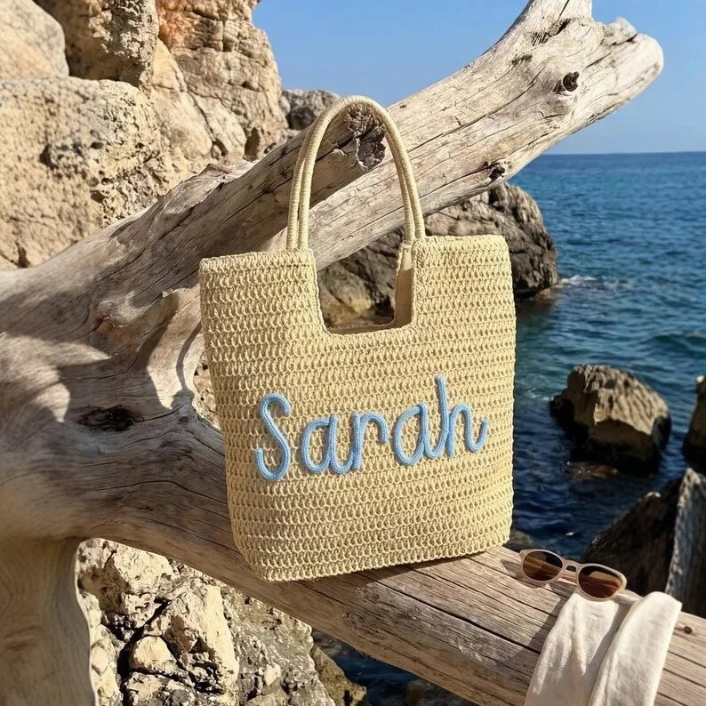 A woven straw tote bag with the name 'Sarah' embroidered in light blue script, resting on a driftwood log by the ocean with rocks and blue sea in the background.