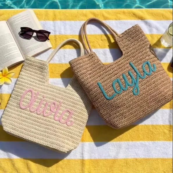 Two woven straw tote bags laid on a yellow-and-white striped beach towel by a pool. One bag is cream with pink embroidered script reading 'Olivia,' and the other is tan with turquoise script reading 'Layla.