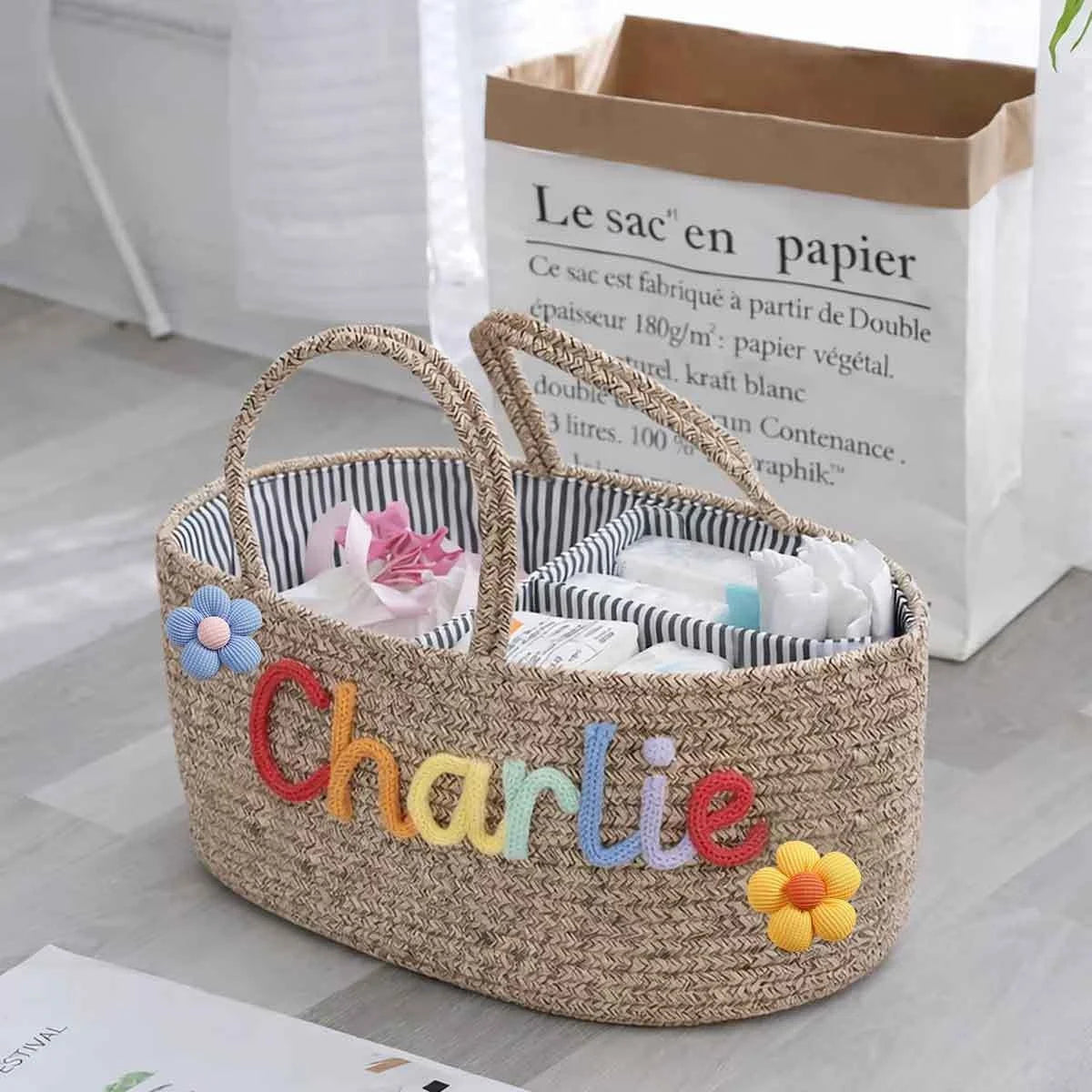 A woven straw storage basket with two handles, personalized with the name 'Charlie' in colorful letters and decorated with flower appliqués. The basket is filled with toiletries and is placed on a light gray floor in front of a white paper bag.