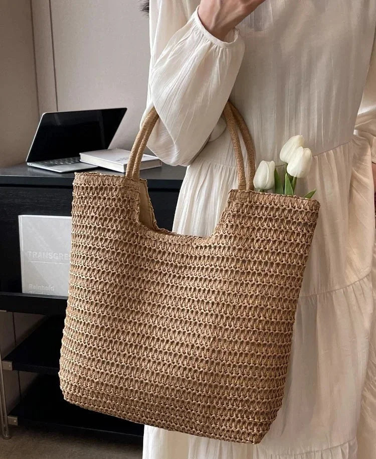 A person in a white dress holds a large woven straw tote bag with two handles. White tulips are tucked into the bag, and a desk with a laptop is visible in the background.