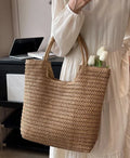 A person in a white dress holds a large woven straw tote bag with two handles. White tulips are tucked into the bag, and a desk with a laptop is visible in the background.