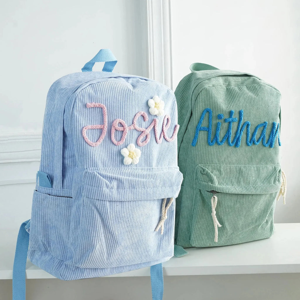 Two personalized corduroy backpacks displayed on a white shelf against a white wall. The backpack on the left is light blue with pink embroidered text reading 'Josie' and white flowers.