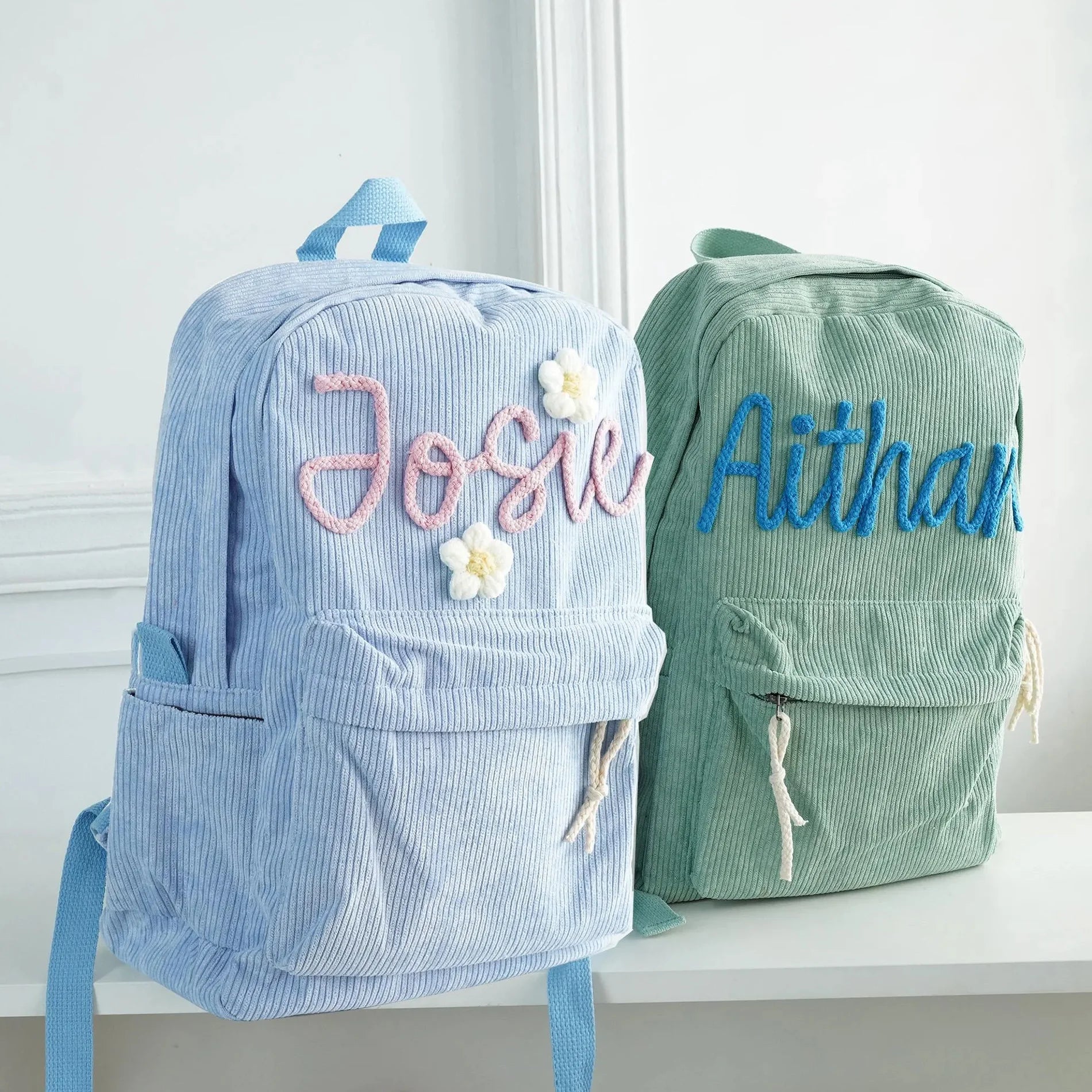 Two personalized corduroy backpacks displayed on a white shelf against a white wall. The backpack on the left is light blue with pink embroidered text reading 'Josie' and white flowers.