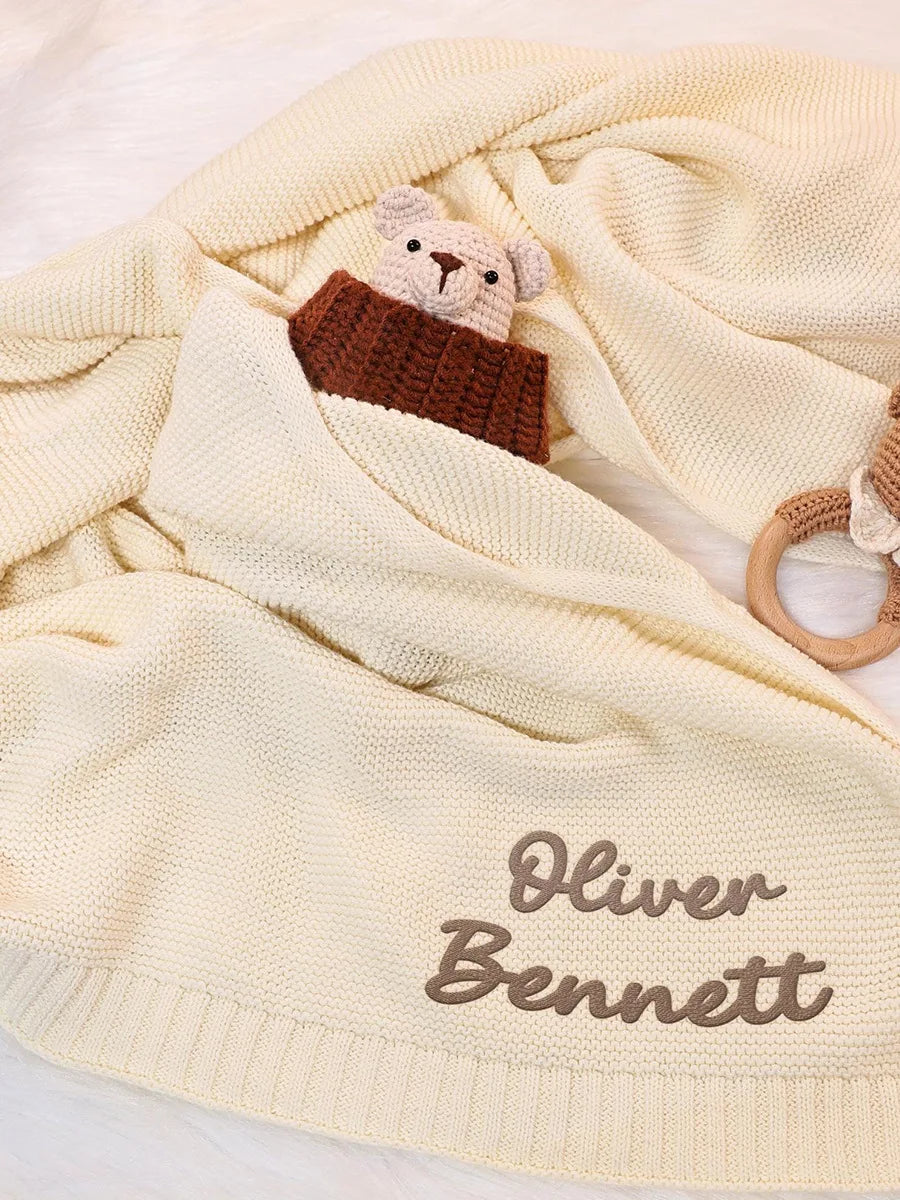 A cream-colored knit baby blanket with personalized embroidery reading 'Oliver Bennett' in brown script, styled with a small plush teddy bear and a wooden teething ring on a soft white background.