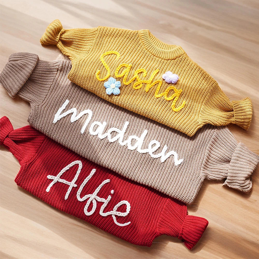 Three personalized ribbed knit baby tights in mustard yellow, taupe, and red, laid flat on a light wood surface. Each pair features a cursive name embroidered across the front: 'Sasha', 'Madden', and 'Alfie', with small decorative flower and cloud embellishments on the top pair.
