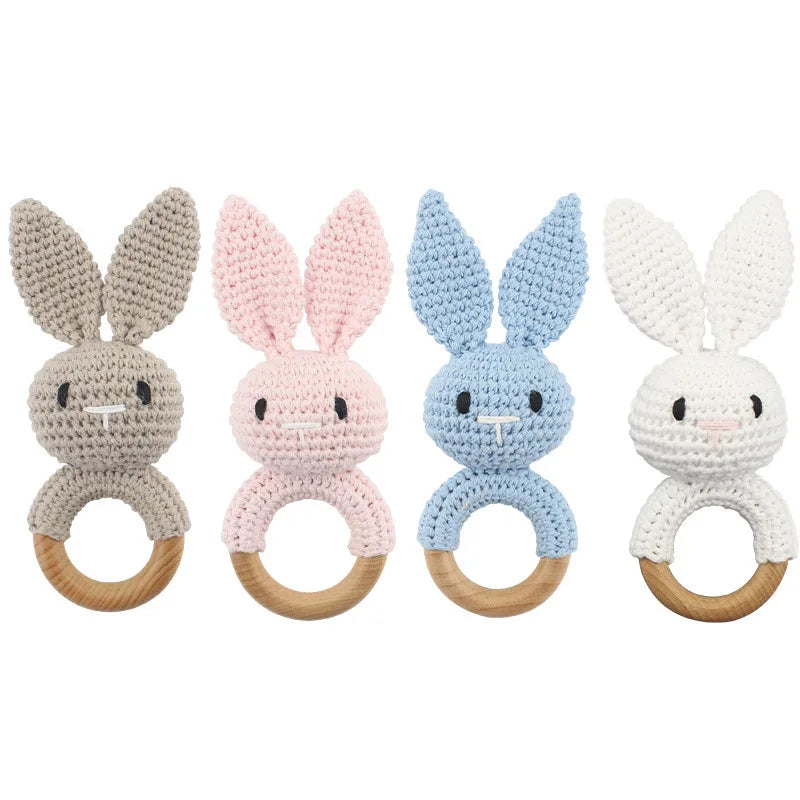 Four handmade crocheted bunny teething rings in beige, pink, light blue, and white, each with a wooden ring handle, shown against a plain white background.