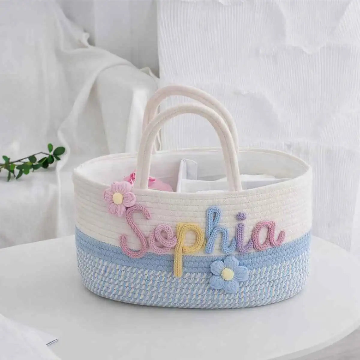 A personalized woven basket bag with the name 'Sophia' embroidered in pastel colors, decorated with two flower appliqués, set on a white surface against a soft white fabric background.