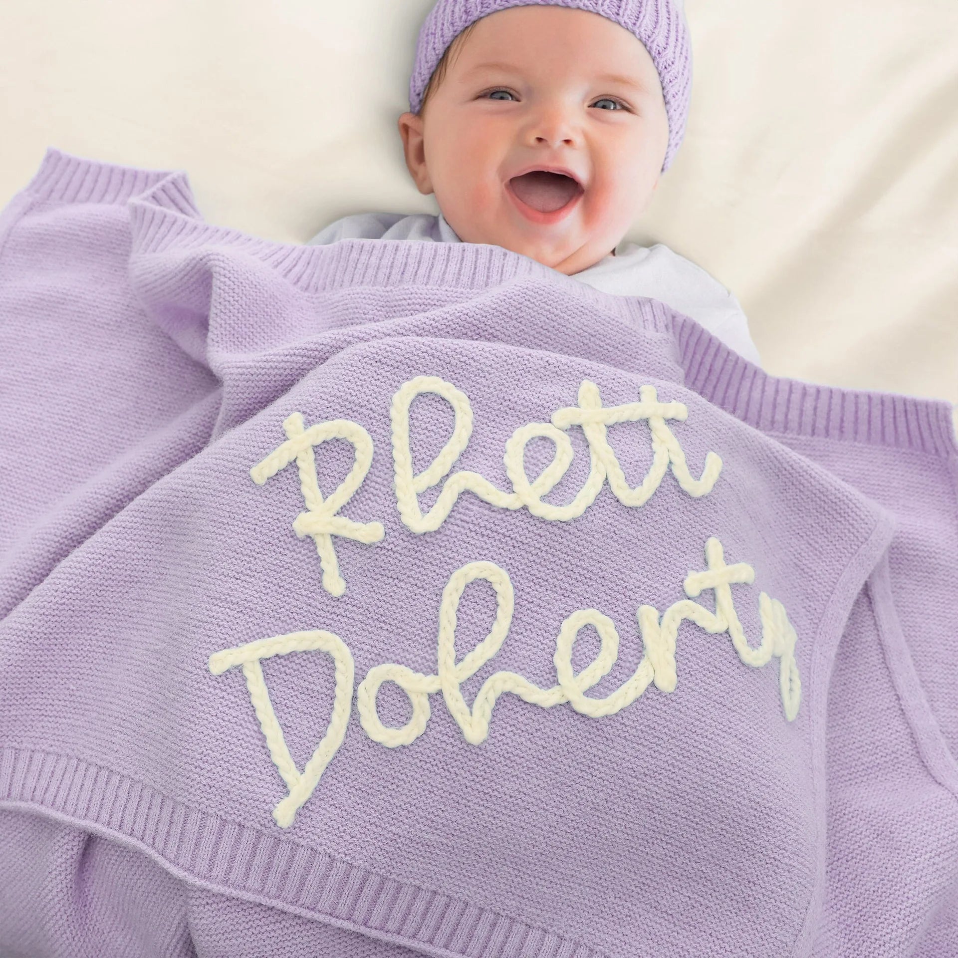 A smiling baby wearing a matching lavender knit beanie and blanket set, with the blanket embroidered with the phrase 'Rett Doherty' in cream lettering.