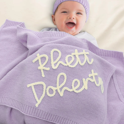 A smiling baby wearing a matching lavender knit beanie and blanket set, with the blanket embroidered with the phrase 'Rett Doherty' in cream lettering.