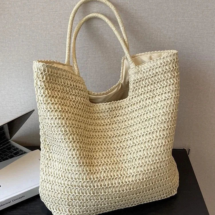 A natural beige woven straw tote bag with two top handles, resting on a dark surface next to a laptop and book against a neutral textured wall.