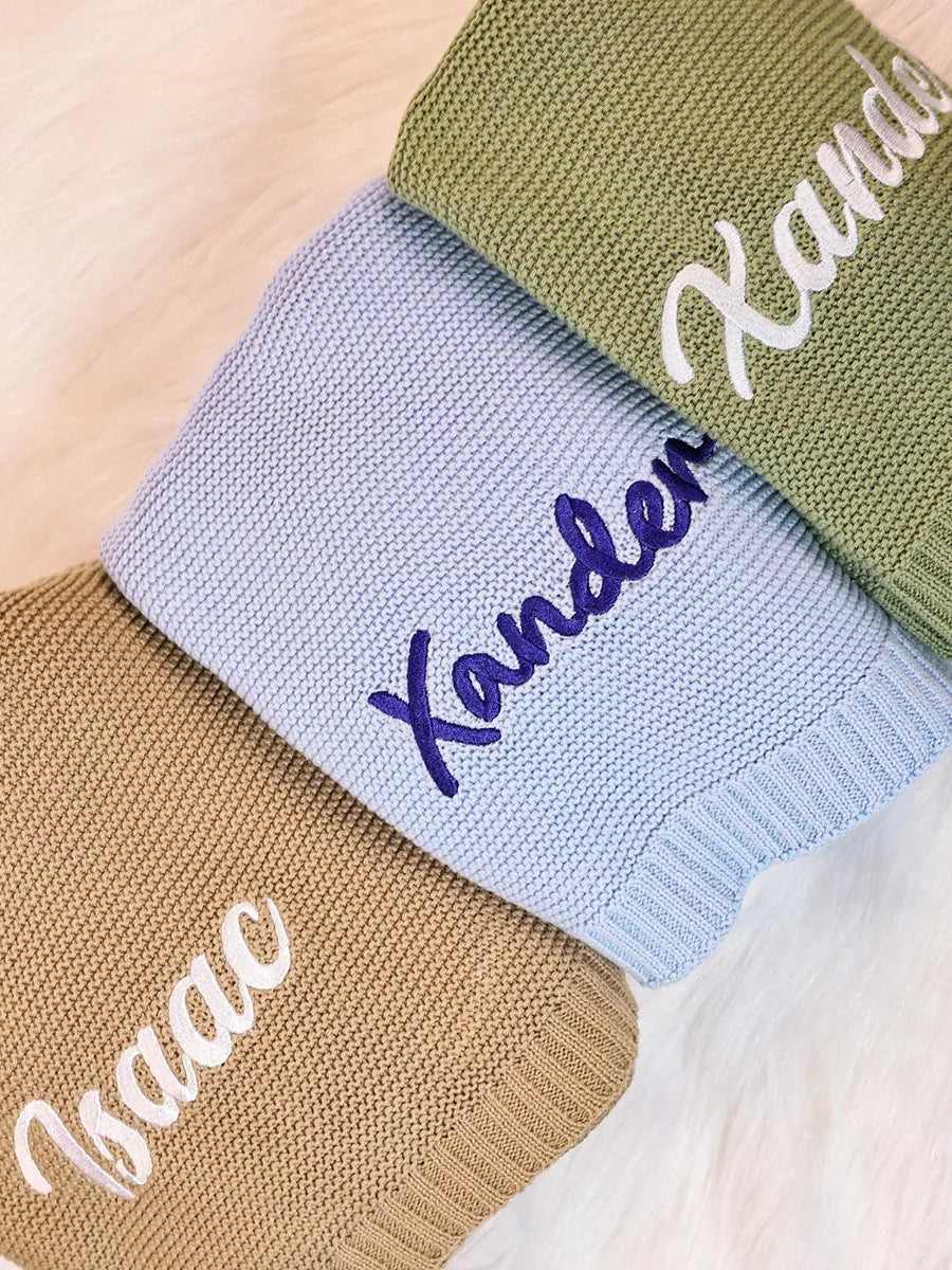 Three knitted socks in beige, light blue, and olive green, each embroidered with a personalized name in script: Isaac, Xander, and Xander. The socks are arranged diagonally on a soft white faux fur surface.
