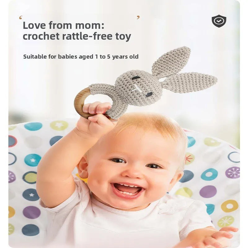 A smiling baby holds a gray crocheted bunny rattle toy with a wooden ring handle. The image includes promotional text reading 'Love from mom: crochet rattle-free toy' and 'Suitable for babies aged 1 to 5 years old' on a soft light background.