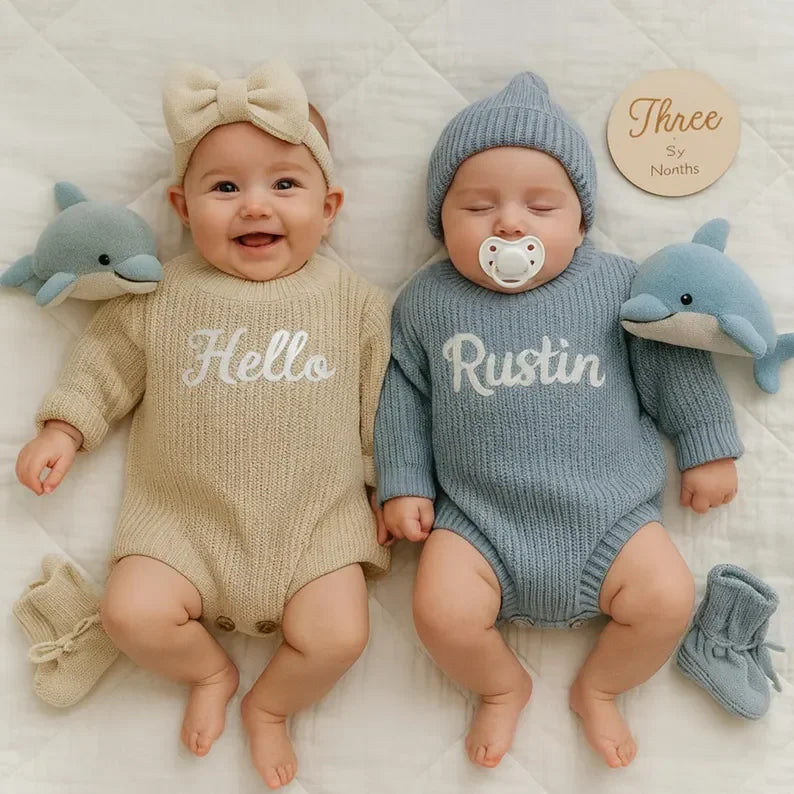 Two babies lying side by side on a white quilted blanket, wearing matching knit baby sets. The baby on the left wears a beige long-sleeve romper with 'Hello' in white script, a beige bow headband, and beige booties.