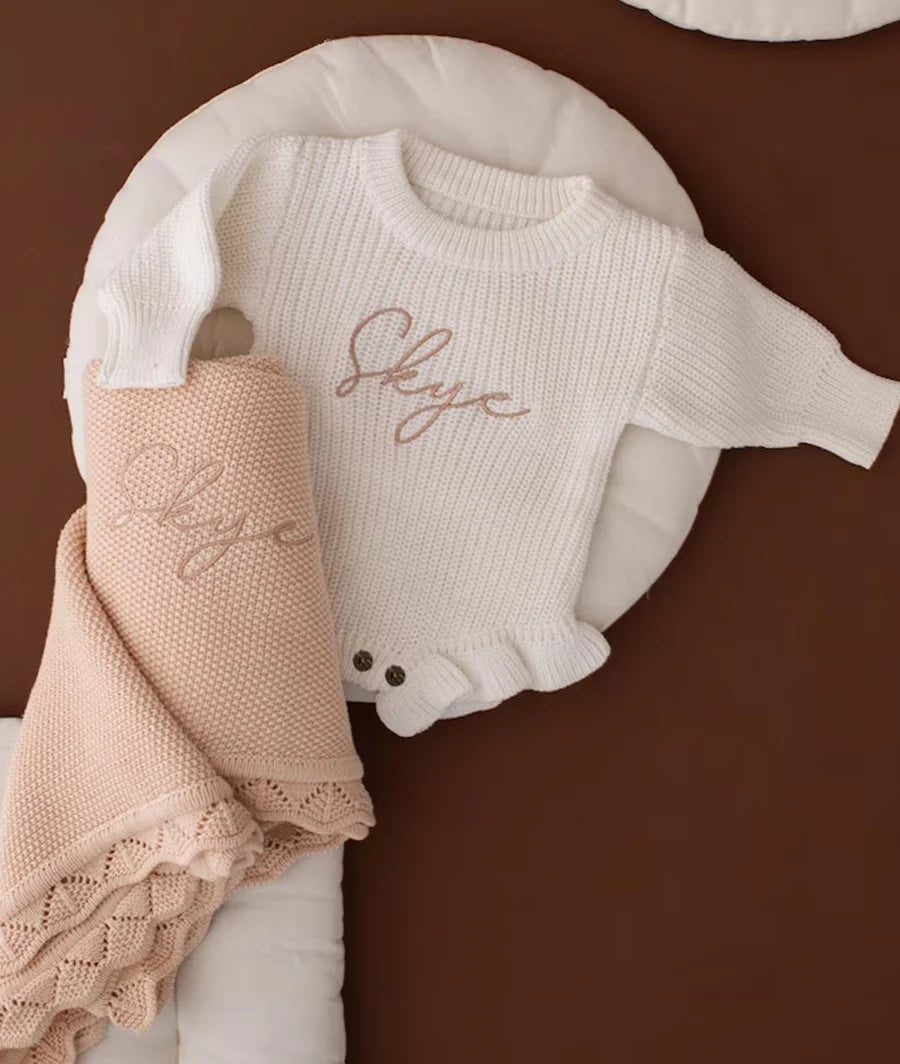 A cream ribbed knit baby sweater with 'Skye' embroidered in rose gold script, styled on a white cushion with a matching cream knit blanket and a blush pink knit throw blanket featuring the same embroidery, all set against a solid dark brown background.