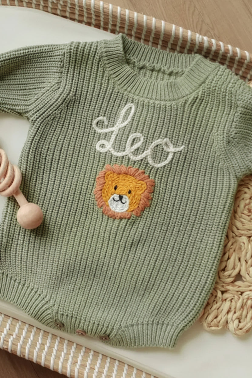 A light sage green ribbed knit baby romper with 'Leo' embroidered in white script and a lion face patch on the chest, styled in a woven basket with a wooden rattle.