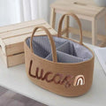 A woven straw organizer basket with two handles, featuring a striped blue and white interior and personalized embroidery reading 'Lucas' with a small rainbow accent on the front.