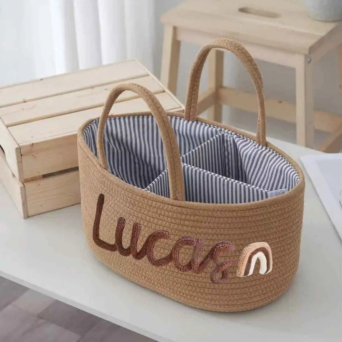 A woven straw organizer basket with two handles, featuring a striped blue and white interior and personalized embroidery reading 'Lucas' with a small rainbow accent on the front.
