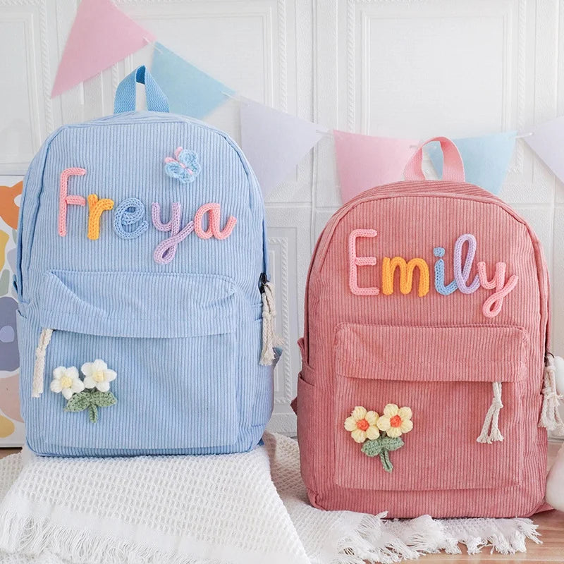Two personalized corduroy backpacks, one light blue with the name 'Freya' and one dusty pink with the name 'Emily', displayed side-by-side against a white paneled wall with pastel bunting.