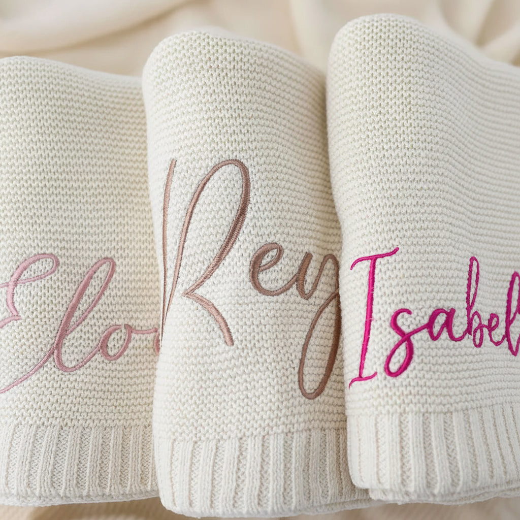 Three rolled cream knit baby blankets with personalized embroidered names in pink, brown, and magenta.
