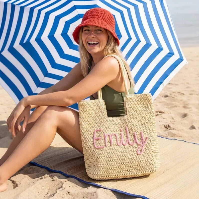 A woman sitting on a beach under a blue and white striped umbrella, wearing a red bucket hat and holding a woven straw tote bag with the name 'Emily' embroidered in pink on the front.