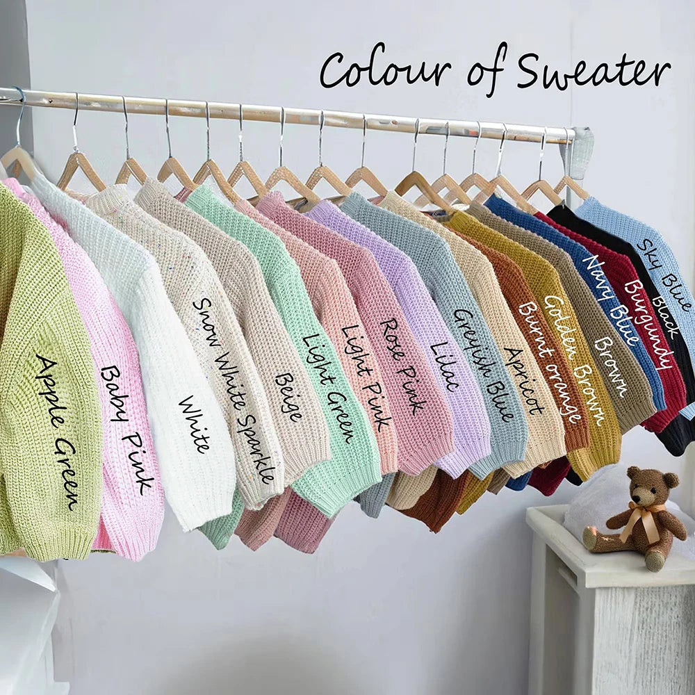 A variety of baby knit sweaters in different colors hanging on a wooden rack, with handwritten color names like Apple Green, Baby Pink, White, Beige, Light Green, Rose Pink, Lilac, Blue, Army Blue, Brown, and Navy Blue. The text 'Colour of Sweater' is written at the top.