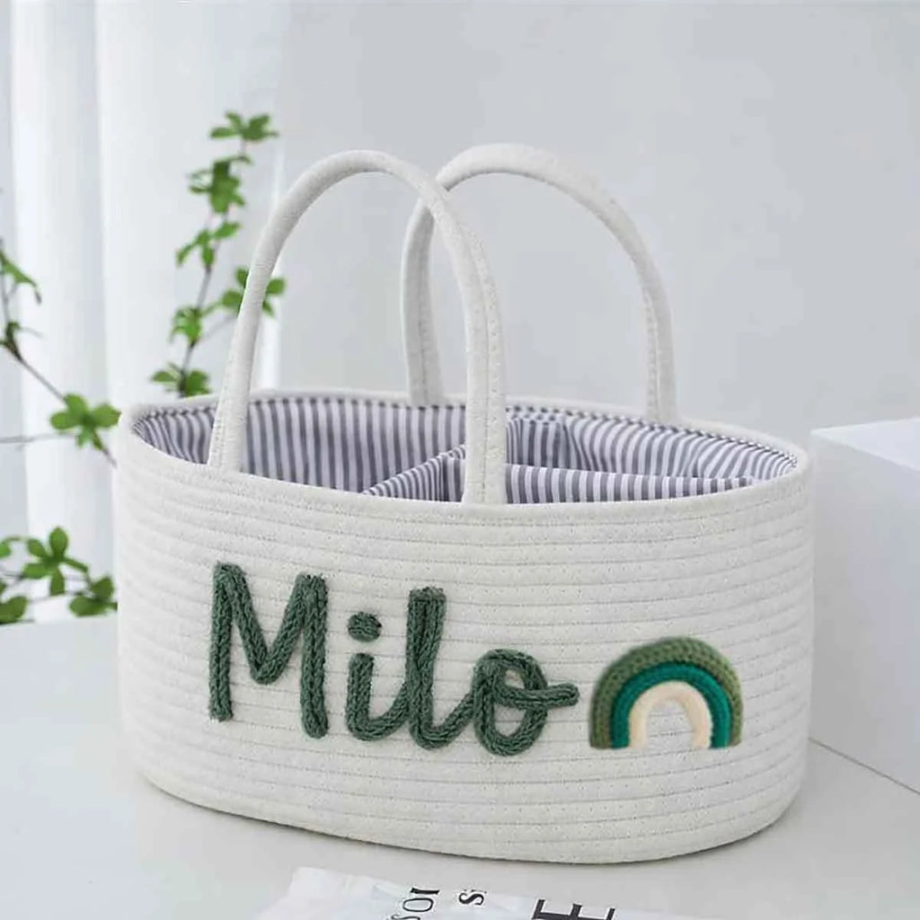 A white woven storage basket with dual top handles, featuring the name 'Milo' embroidered in dark green thread and a small rainbow motif on the front. The basket is lined with a blue and white striped fabric and is set on a light surface with a blurred indoor background.