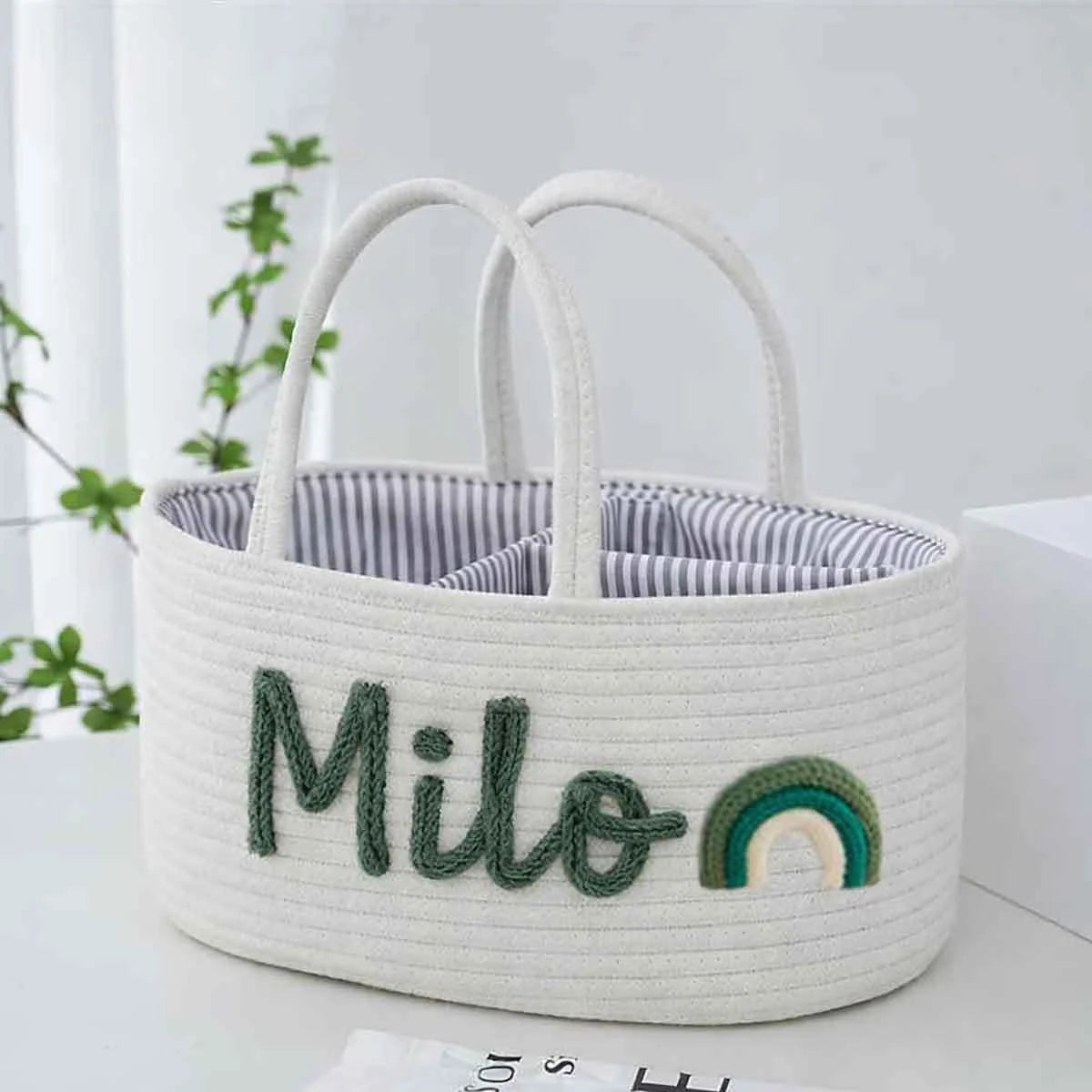 A white woven storage basket with dual top handles, featuring the name 'Milo' embroidered in dark green thread and a small rainbow motif on the front. The basket is lined with a blue and white striped fabric and is set on a light surface with a blurred indoor background.