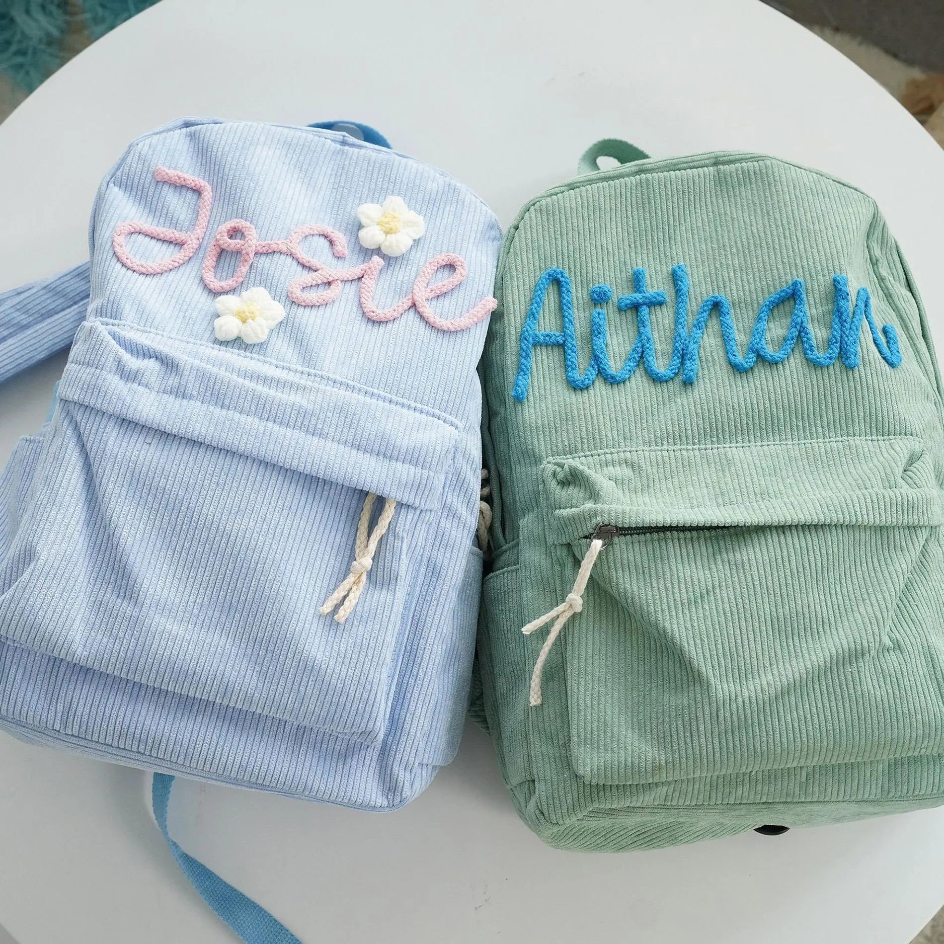 Two backpacks, one blue and one green, with embroidered names and flower designs.
