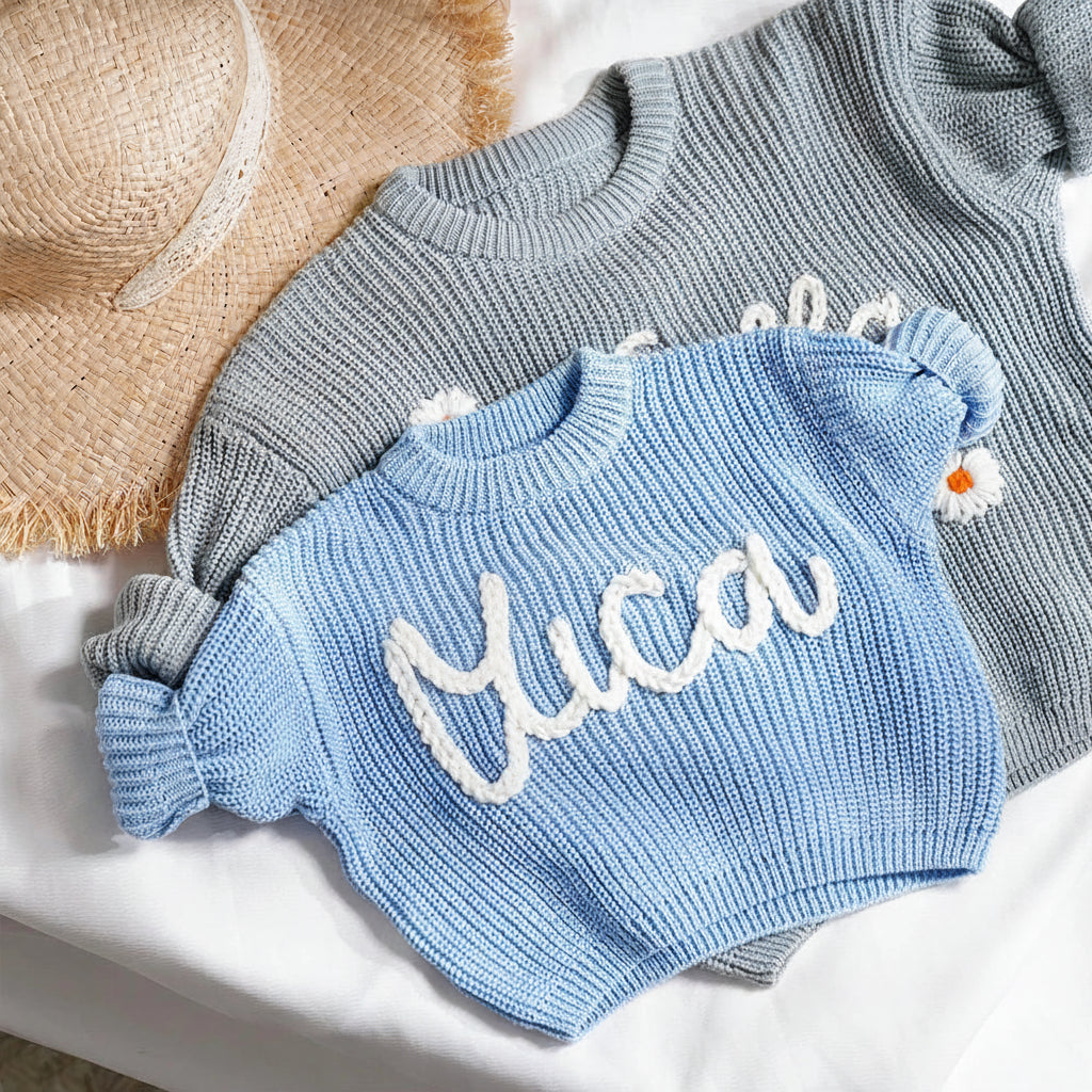 A light blue knit sweater with 'luca' written in white script and a small daisy detail, styled on top of a gray knit sweater and a straw hat on a white fabric background.